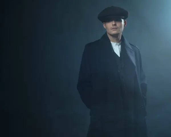 Mężczyzna w kaszkiecie i płaszczu, z charakterystycznym wąsem, emanuje chłodnym spojrzeniem w stylu Peaky Blinders.