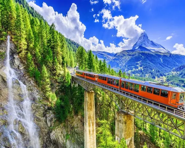 An orange train crosses a bridge over a waterfall, on its way from Zurich to Andermatt. Lush green trees and mountains surround the scenic route.