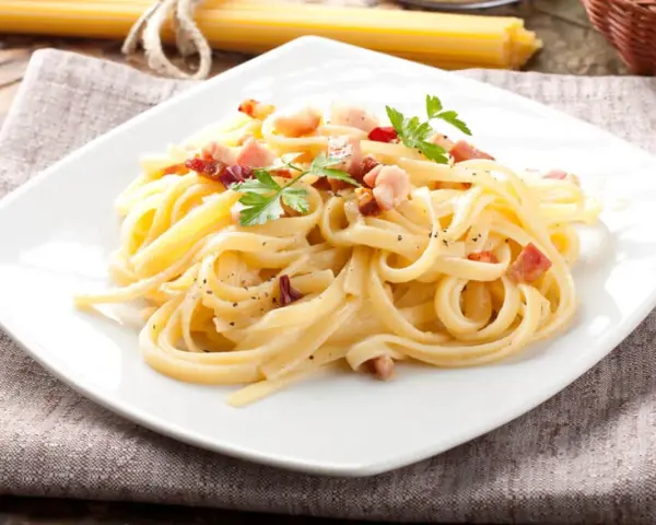 Pyszne spaghetti carbonara z boczkiem i natką pietruszki na białym talerzu.