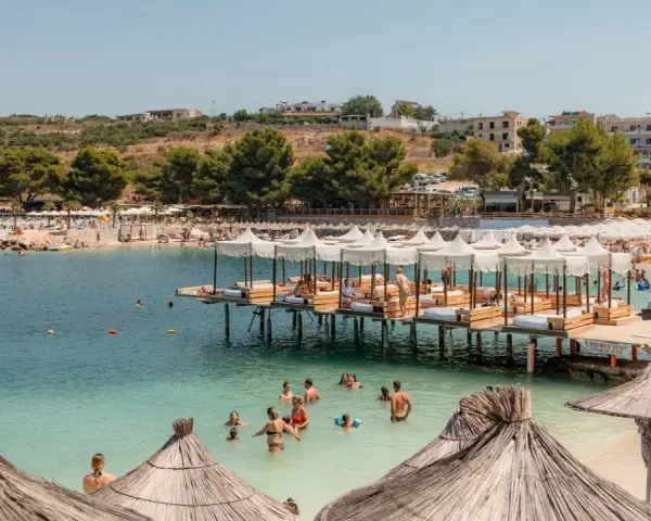 Słoneczny dzień na Ksamil Beach. Ludzie kąpią się w turkusowej wodzie, relaksują się na leżakach pod białymi parasolami i na drewnianych pomostach.