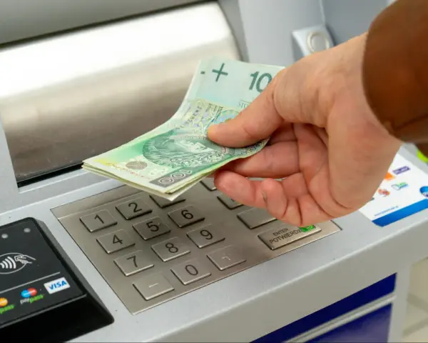 Dłoń wkłada banknoty 100 zł do bankomatu. Zastanawiasz się, ile można wpłacić na konto?