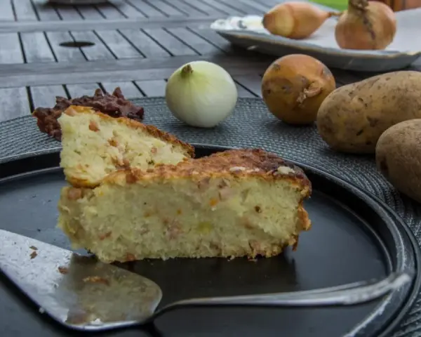 Babka ziemniaczana przepis babci: złocisty placek z ziemniaków, jajek i boczku, obok surowe ziemniaki, cebula i jajko.