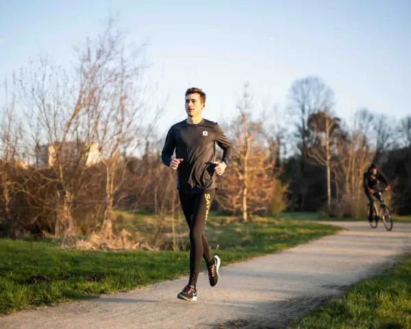 Un jeune homme s'entraîne à la course à pied sur un chemin ensoleillé, entouré d'arbres dénudés. Un cycliste passe en arrière-plan.