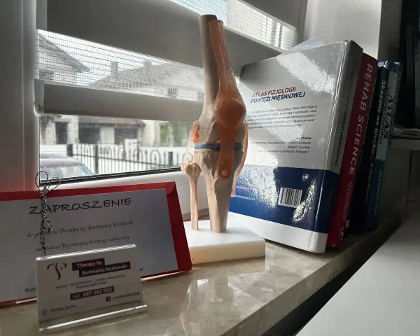 Model stawu kolanowego obok zaproszenia na terapię od Bartłomieja Kozłowskiego i książek o fizjoterapii.