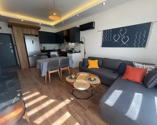 Nowoczesny salon z aneksem kuchennym. Wygodna kanapa, stolik kawowy, stół z krzesłami. W tle lodówka i sprzęty kuchenne. Idealne miejsce na relaks, z myślą o komforcie gości.