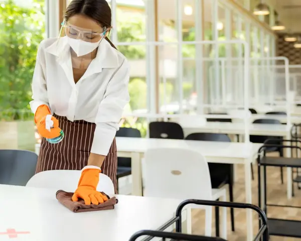 Employée portant masque et visière nettoie une table, respectant la réglementation hygiène cantine scolaire. Des séparateurs sont installés.
