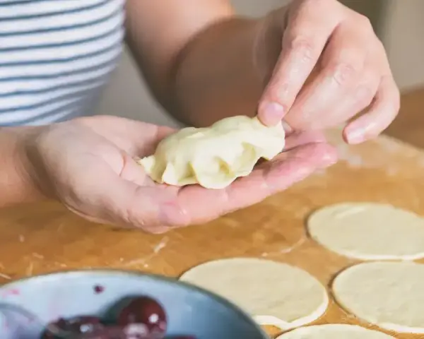 Ręce formują pierogi ruskie na drewnianej desce. W tle miska z wiśniami.