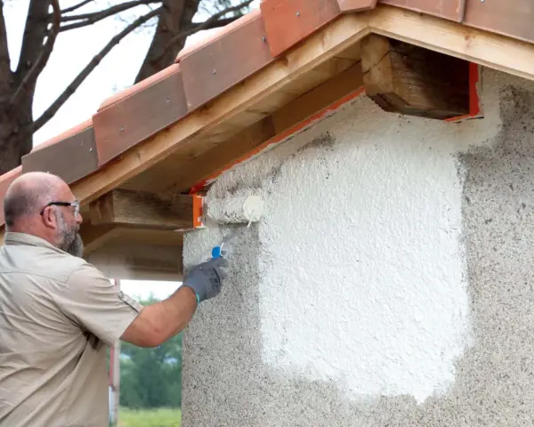 Un homme applique une peinture sur mur béton extérieur avec un rouleau. La rénovation progresse sous le toit de tuiles.