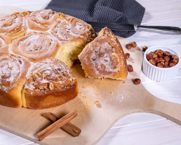 Ein köstliches Zimtschnecken Rezept: goldbraune, mit Nüssen und Puderzucker bestreute Schnecken auf einem Holzbrett, daneben Zimtstangen und Haselnüsse.