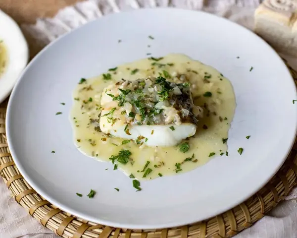 Un delicioso bacalao en salsa verde, con perejil fresco picado y trozos de pan al lado.