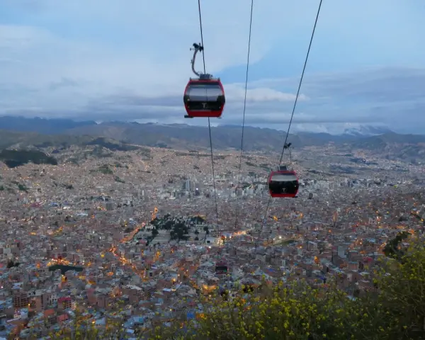 Red la paz gondola cabins glide over a sprawling city at dusk, with lights twinkling below and mountains in the distance.