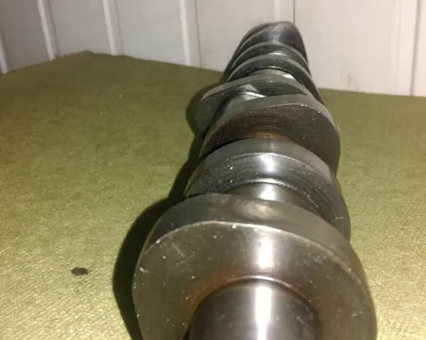 Metalowy, wytarty wałek rozrządu leży na zielonym materiale. Widać ślady zużycia na jego krzywkach.