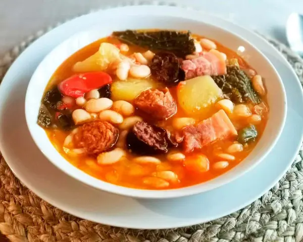 Un reconfortante cocido montañés, rebosante de alubias blancas, patatas, chorizo, morcilla y tocino, servido en un cuenco blanco.