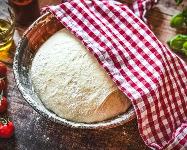 Frischer Pizzateig ruht in einer Schüssel, bedeckt mit einem rot-weiß karierten Tuch. Zutaten für ein Pizzateig Rezept wie Tomaten und Öl liegen bereit.