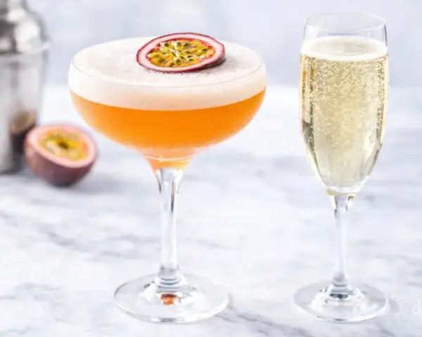 Pomarańczowy drink z pianką i marakują, obok kieliszek szampana. Idealny white lady drink na letnie popołudnie.