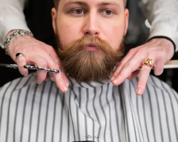 Fryzjer precyzyjnie modeluje brodę w szpic, używając nożyczek i grzebienia.
