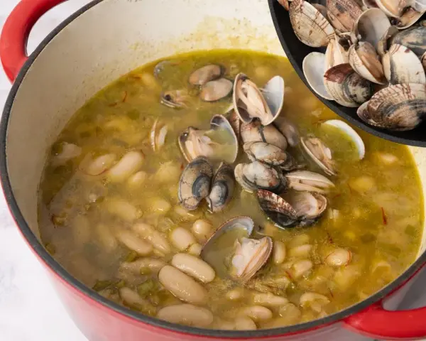 Alubias cremosas con almejas en una olla roja, añadiendo más almejas de una sartén.