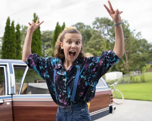 Millie Bobby Brown w kolorowej koszuli i szelkach, radośnie pozuje przy samochodzie. Jej filmy, seriale i programy to gwarancja emocji!