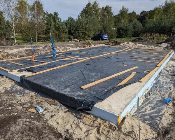 Gotowy fundament pod dom szkieletowy, z czarną folią i drewnianymi belkami, czeka na dalsze prace budowlane.