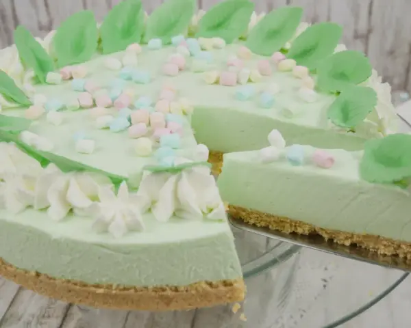 Eine helle, cremige Philadelphia Torte Waldmeister mit bunten Marshmallows und grünen Blättern verziert. Ein Stück wird gerade herausgeschnitten.