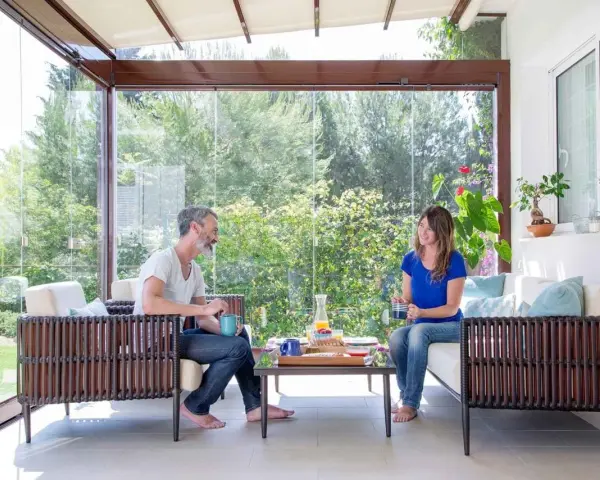Pareja disfrutando de un café en un **porche acristalado** con vistas a un jardín exuberante.