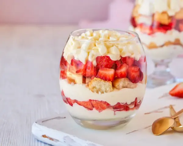Postre en vaso con capas de crema, fresas y bizcocho, cubierto con trozos de chocolate blanco.