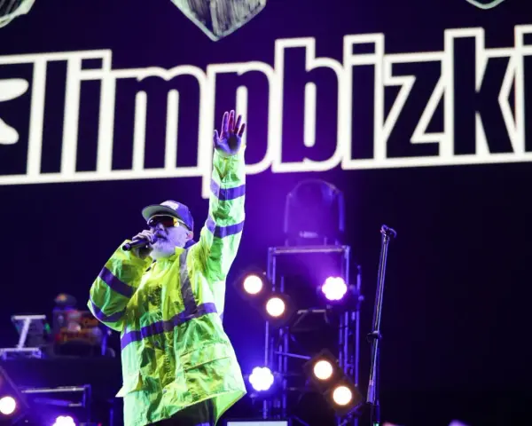 Fred Durst z Limp Bizkit na koncercie, w neonowej kurtce, z ręką w górze. Tło to logo zespołu.