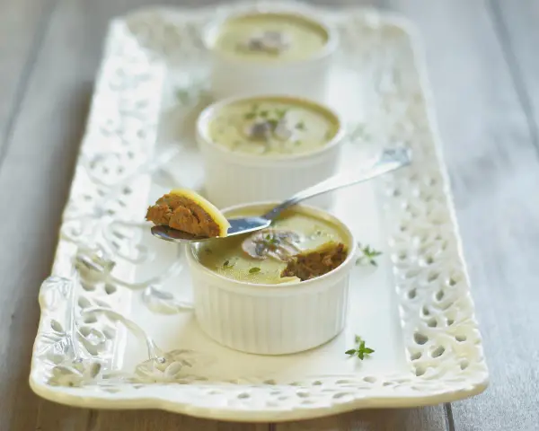 Trois ramequins de foie de volaille, ornés de champignons et de persil, sur un plateau ajouré. Une cuillère présente une bouchée de ce délice.
