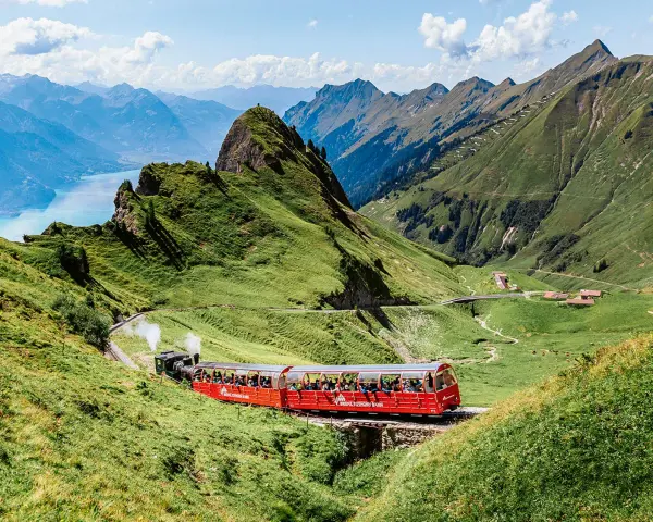 A red Brienz Switzerland train winds through lush green mountains, with a stunning blue lake in the distance.