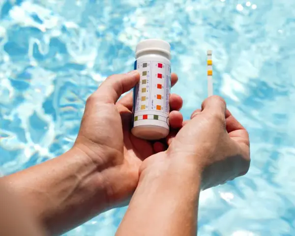 Hands hold a pool test strip and bottle, showing how to make pool water clear by checking chemical levels.
