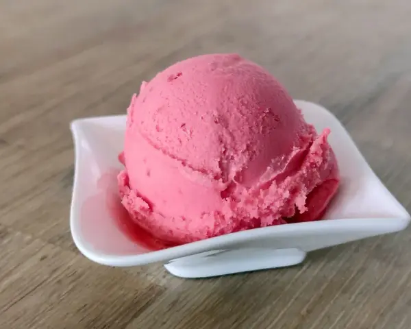 Ein Löffel cremiges Himbeersorbet in einer weißen Schale auf Holzhintergrund.