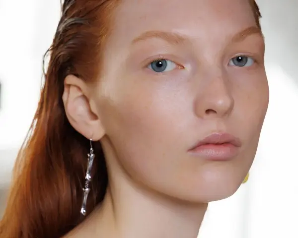 Primo piano di una giovane donna con capelli rossi e occhi azzurri, un neo della bellezza sul viso. Indossa un orecchino pendente.