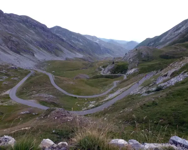 Strada tortuosa che sale verso il colle dei morti, tra montagne verdi e rocciose. Due ciclisti affrontano le curve.