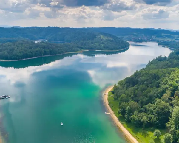 Idealne wakacje w górach nad jeziorem. Zielone wzgórza, turkusowa woda i spokojne żaglówki tworzą malowniczy krajobraz.