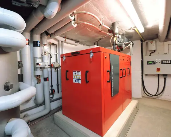 Rotes Blockheizkraftwerk im Keller eines Mehrfamilienhauses, umgeben von Rohren und einer Steuereinheit.