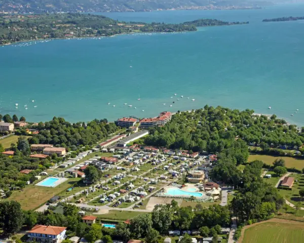 Widok z lotu ptaka na camping Baia Verde nad turkusowym jeziorem, z basenami i licznymi namiotami.
