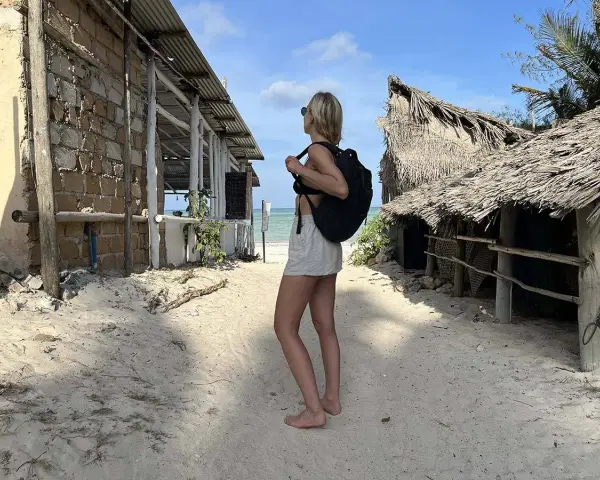 Kobieta z plecakiem na piaszczystej plaży, zastanawia się co zabrać na Zanzibar. W tle chatki z palmowymi dachami i budynek z kamienia.