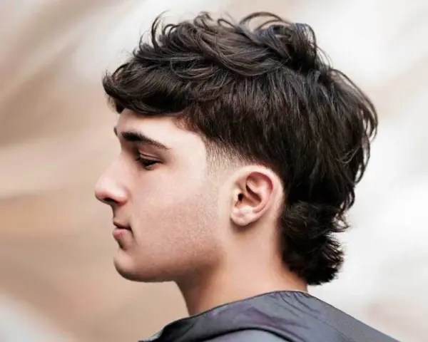 Młody mężczyzna z kręconymi włosami i modną fryzurą mullet. Krótsze boki i dłuższe włosy z tyłu nadają mu charakteru.