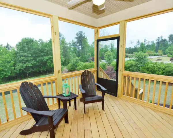 A screened-in porch with two Adirondack chairs, a small table, and a green lantern. The porch offers a view of lush trees and a suburban landscape.