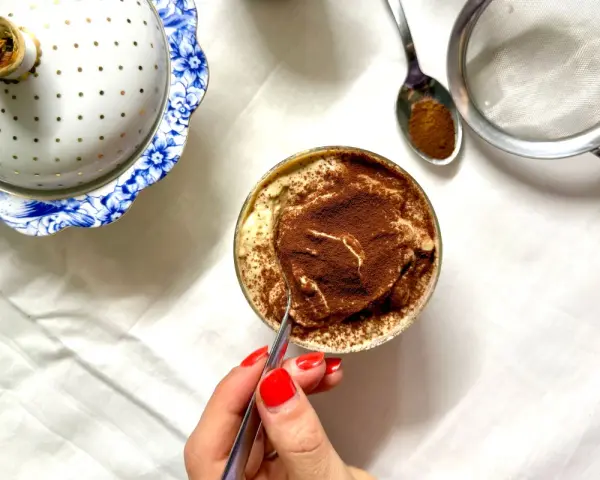 Deliziosa crema al caffè in bicchiere, cosparsa di cacao. Accanto, un elegante contenitore bianco e oro e un cucchiaino con cacao.