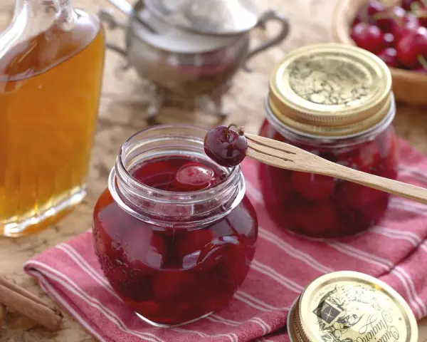 Frascos de mermelada de cerezas casera, una botella de licor y canela en rama. Una cereza en un tenedor de madera lista para probar.
