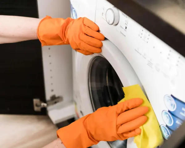 Hände in orangen Handschuhen reinigen die Waschmaschine. Ein gelber Lappen wischt über die Oberfläche, um sie sauber zu halten.
