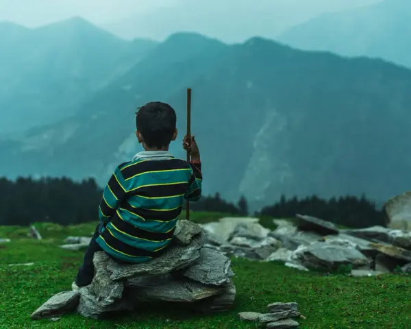 Junge sitzt auf Steinen, blickt in nebelverhangene Berge. Ein Stock in der Hand, wie für einen meditationstext zum vorlesen.