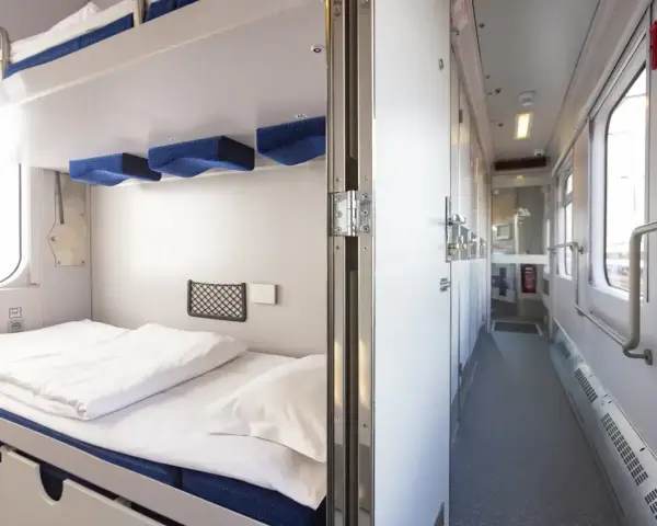 Wnętrze wagonu sypialnego PKP 3 osobowego. Przytulne łóżka, czyste pościel i korytarz z oknami.