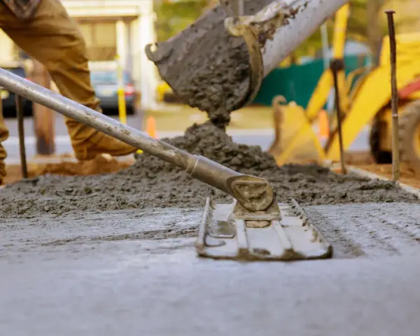 Pracownik rozprowadza świeży beton c12/15 za pomocą łaty. W tle widać koparkę i plac budowy.