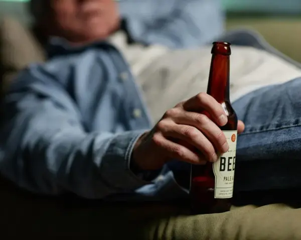 Mężczyzna w dżinsach i koszuli trzyma butelkę piwa, leżąc na kanapie. Może to sugerować spalanie alkoholu podczas snu.