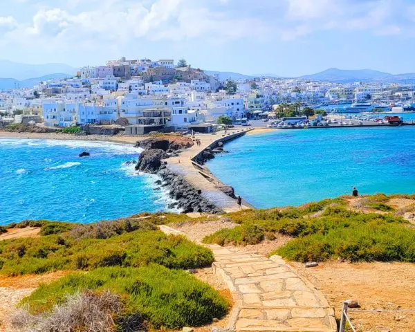 A stone path leads to a causeway connecting to the whitewashed town of Naxos, Greece, with vibrant blue waters lapping at the shore.