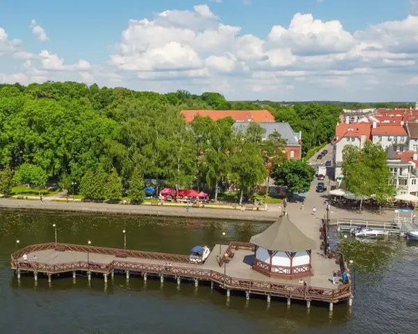 Malownicze mazury miejscowości nad jeziorem. Drewniane molo z altaną, zielone drzewa i urokliwe kamienice tworzą letni klimat.