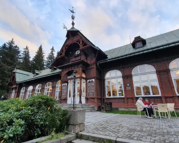 Drewniany pawilon z zegarem i karłowata studzienka przed wejściem. Na zewnątrz siedzą ludzie przy stoliku.