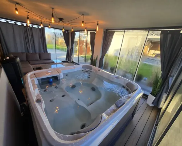 Relaks w jacuzzi pod ciepłym światłem lamp w przeszklonym tarasie zimowym. Obok wygodna sofa i stolik.